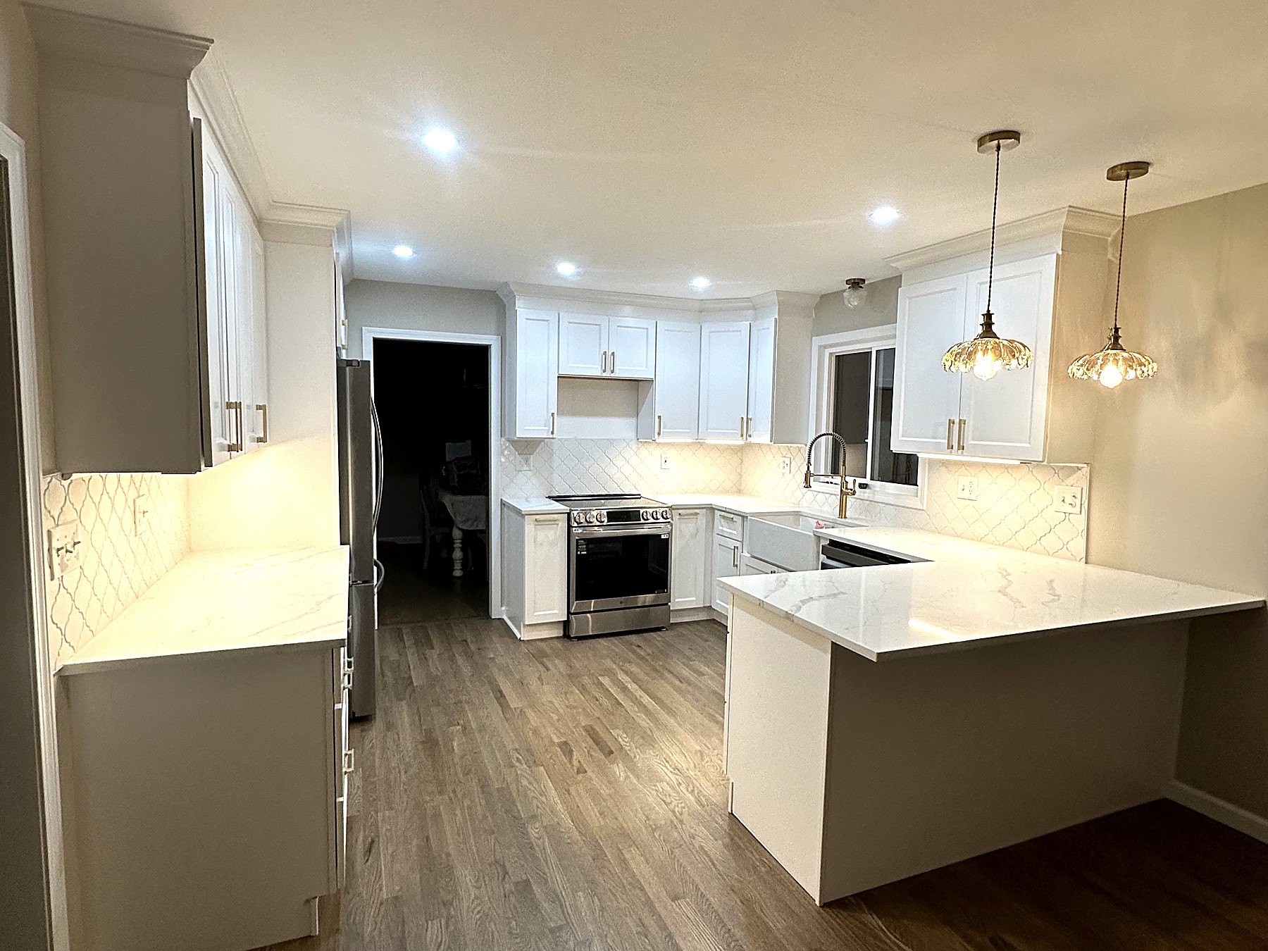 Renovated kitchen in Terryville Connecticut with white shaker cabinetry and crown molding, marble-look quartz countertops, arabesque tile backsplash, farmhouse sink with brass faucet, stainless gas range, brass pendant lights over a peninsula bar, and red oak hardwood flooring