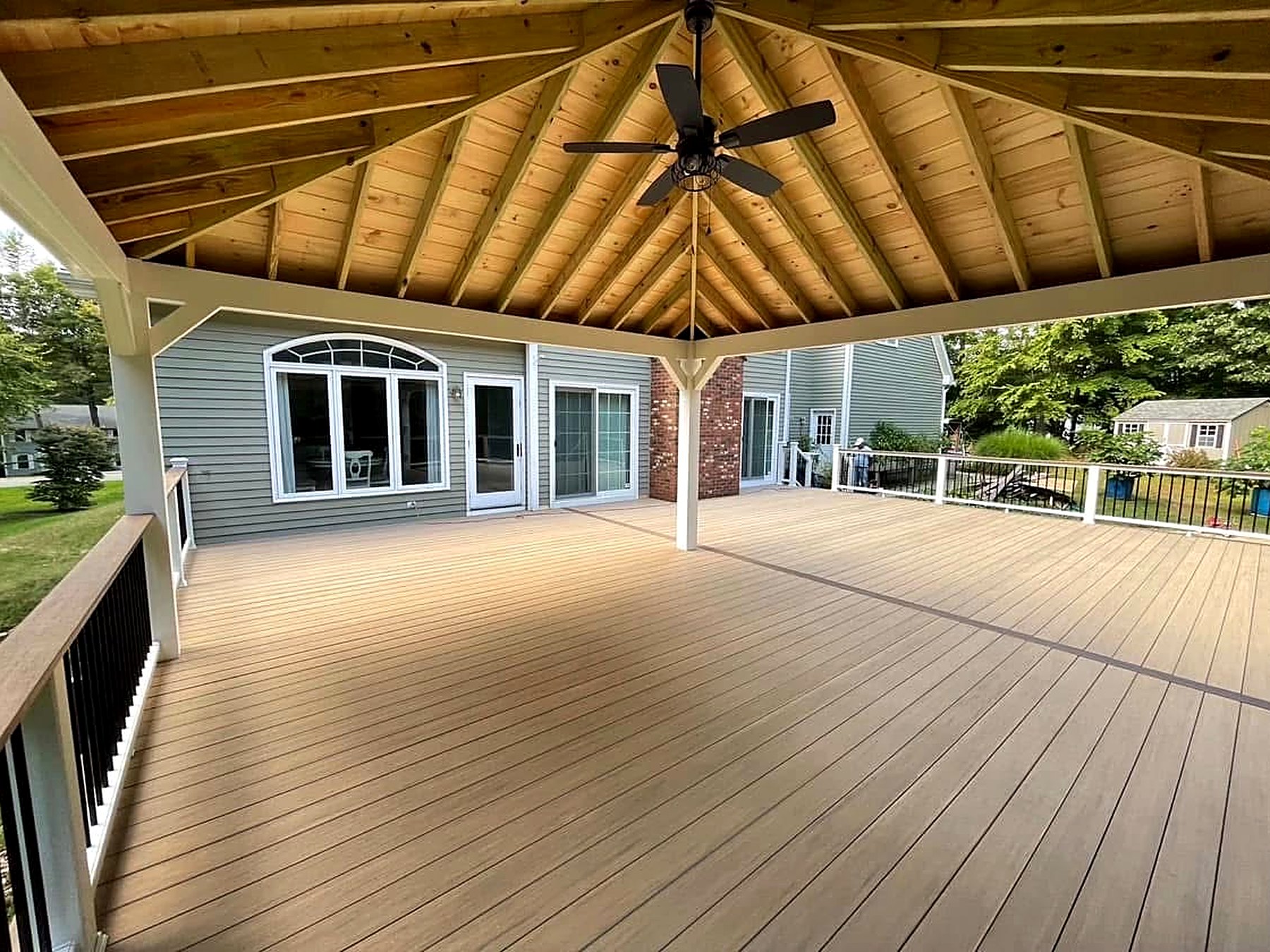 Rebuilt composite deck in Darien Connecticut under a new open-gable covered porch with a cathedral cedar tongue-and-groove ceiling, exposed rafters, white wrapped posts and beams, black ceiling fan, and white railing with black aluminum balusters