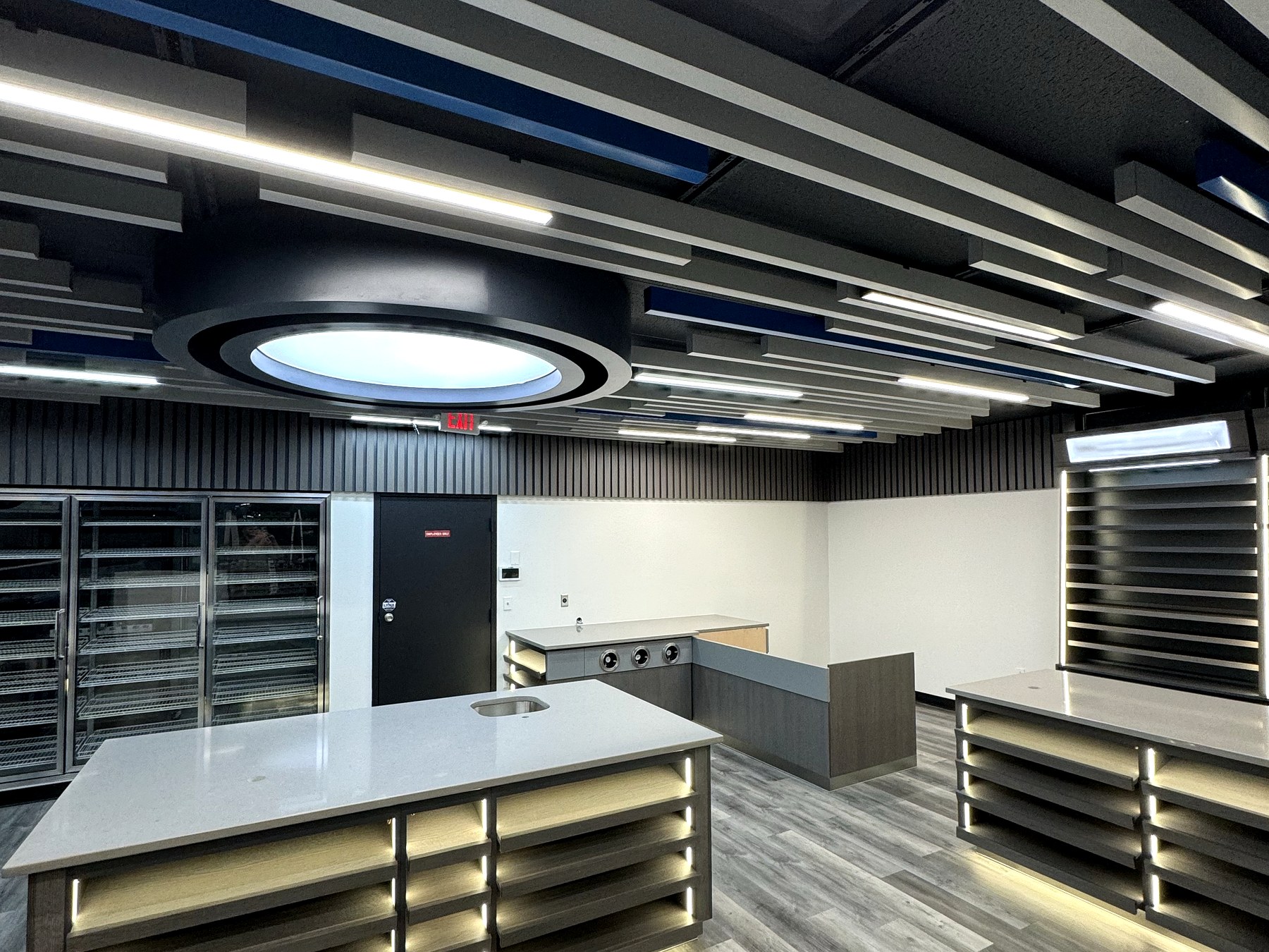 Renovated gas station convenience store sales floor in Northford Connecticut with a black acoustic baffle ceiling with navy accents and linear LED strips, a circular illuminated ceiling feature, a glass-door reach-in cooler line, black slat wall panels, LED-lit island gondola displays with light quartz tops, a coffee and beverage service counter, and grey wide-plank commercial vinyl plank flooring