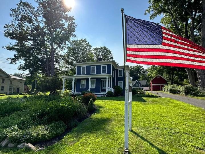 Historic colonial home in Woodbury Connecticut after a full exterior renovation, with deep navy siding, white trim, a restored full-width front porch, and a red barn on the property