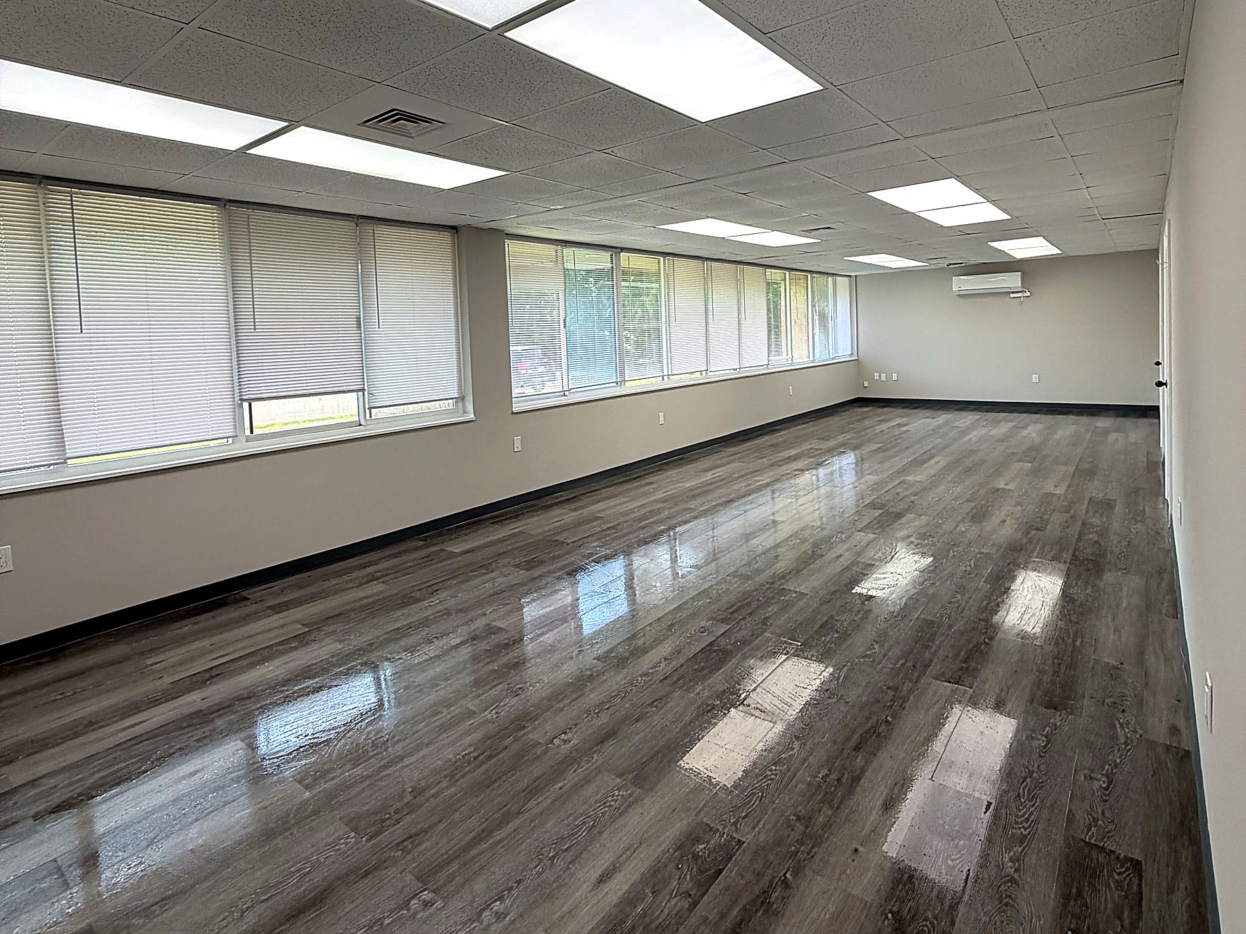 Commercial employee break room in Ansonia Connecticut after a full build-out, with grey wide-plank vinyl flooring, black vinyl cove base, warm grey painted walls, a new suspended acoustic tile ceiling with recessed LED flat panels, a full wall of windows with new blinds, and a wall-mounted ductless mini-split