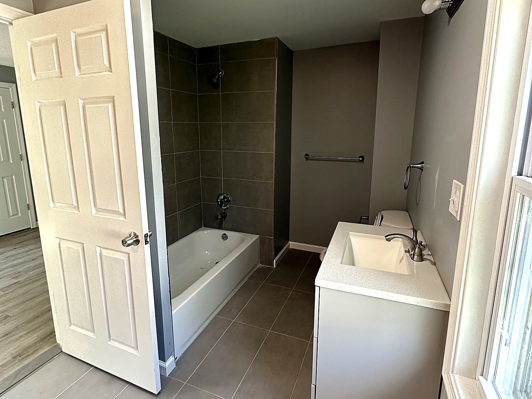 Remodeled bathroom in Watertown Connecticut with large-format grey porcelain floor tile, a charcoal tile tub and shower surround with chrome trim, a white soaking tub, a white vanity with a solid-surface top and chrome faucet, warm grey walls, chrome vanity lighting, and a new ceiling exhaust fan and light